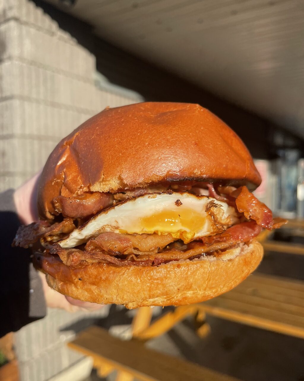 A bacon and egg brioche bap held up in front of wooden picnic tables.
