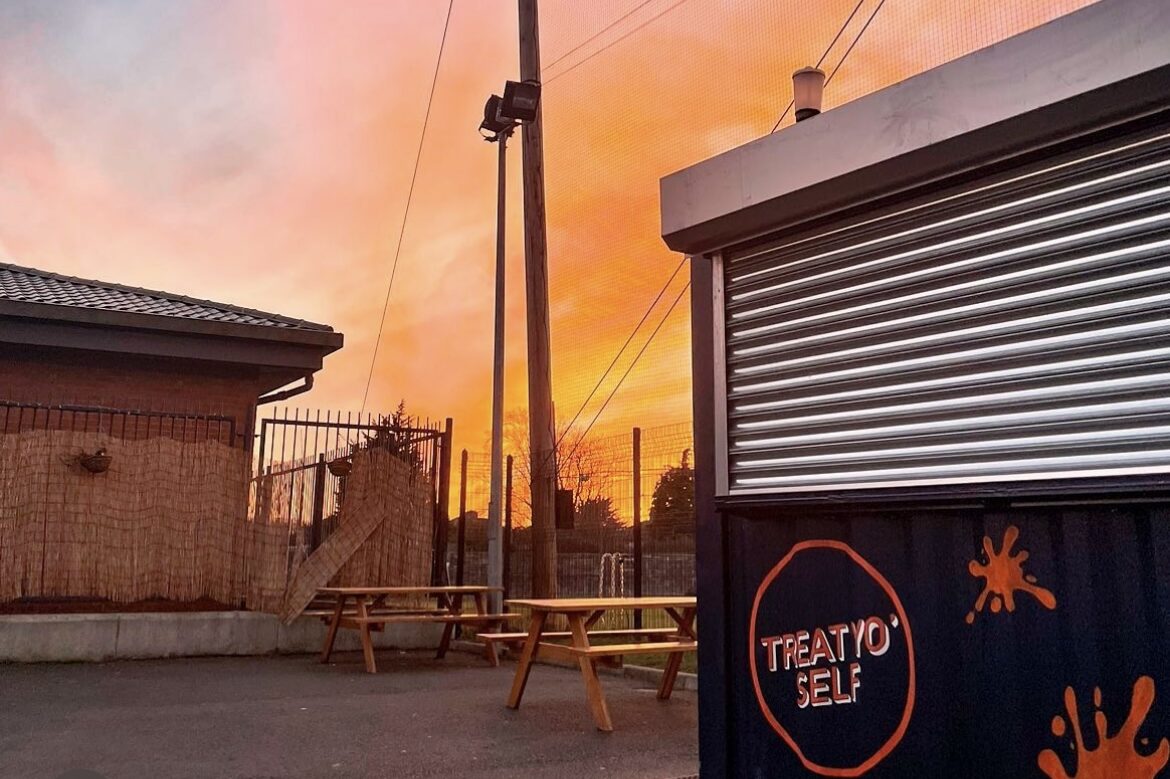 The Treat Yo'Self kiosk at sunset: a navy container with its roller shutter down, orange splats, and picnic tables beside it.