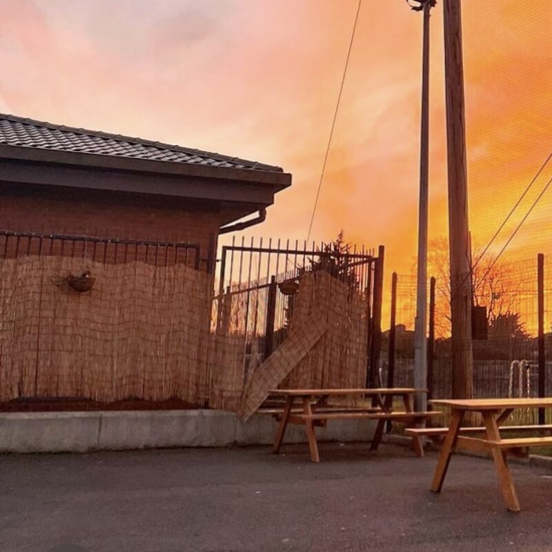Wooden picnic tables on tarmac under an orange evening sky.
