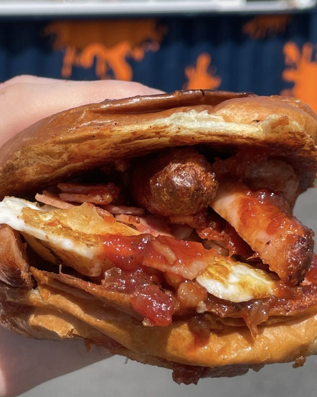 A hand holding a bap filled with sausage, bacon, egg and beans in front of the navy container with orange splats.